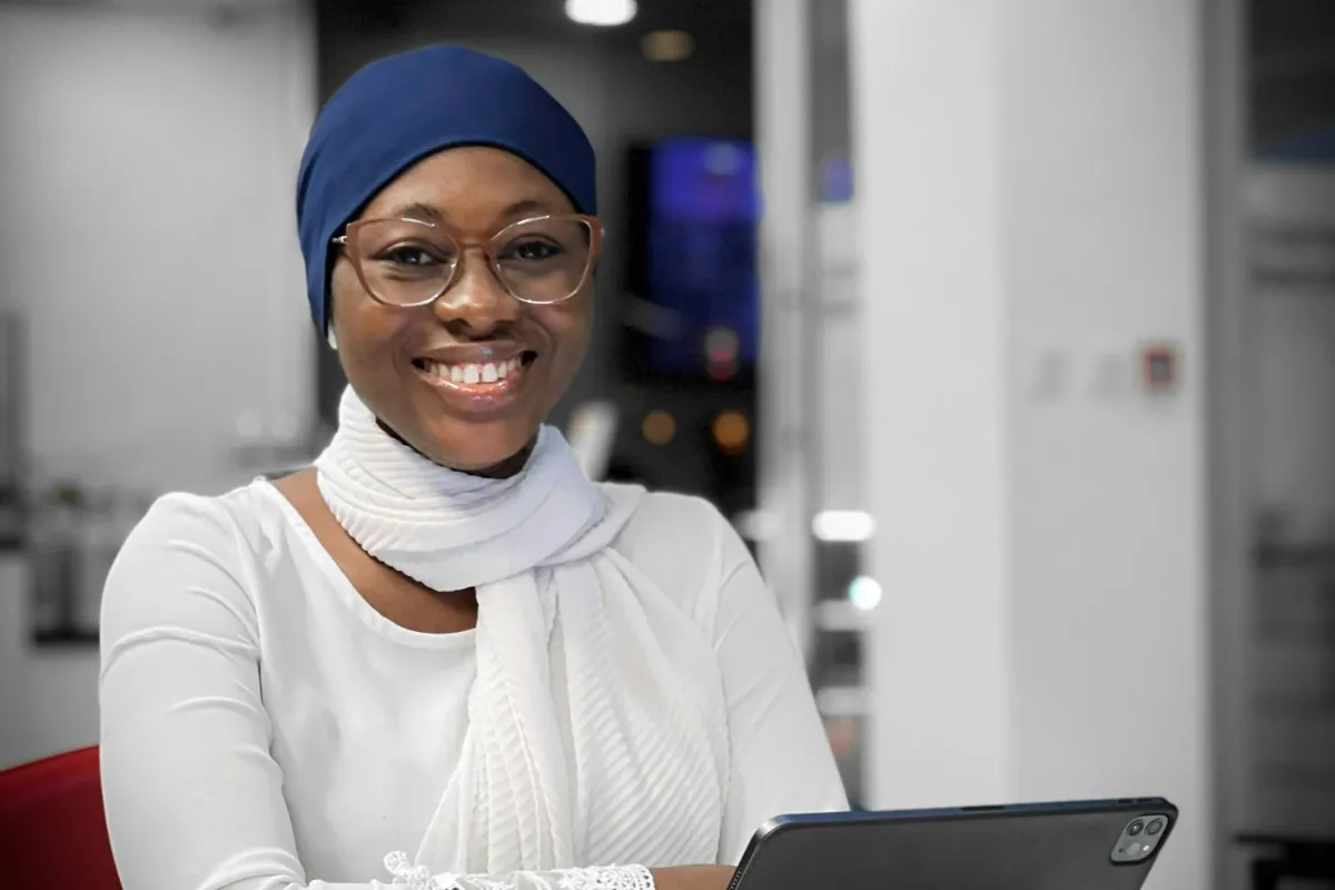 A smiling woman in glasses and a headscarf sitting at a table with a tablet